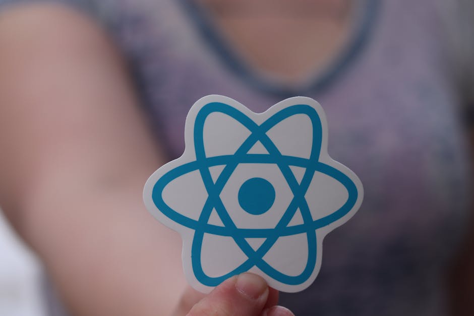 A person holding a sticker featuring a blue and white atom symbol, with a blurred background showing a person wearing a grey top and a room with neutral-colored walls, indicating clean and well-mainta