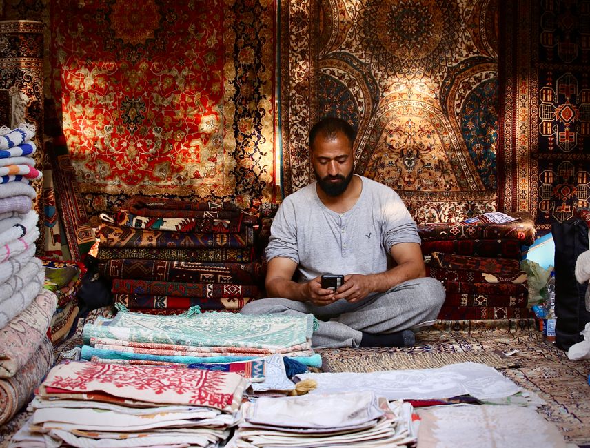 A display of various rolled carpets in a shop or storage area, arranged vertically against a wall. The carpets feature intricate patterns in shades of red, beige, blue, and brown, and are made from ma
